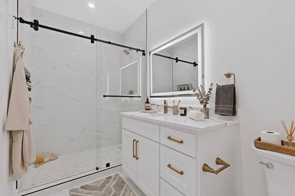7503 Robin Road, Dallas, TX 75209 Walk-in shower with frameless glass sliding enclosure, stone-finish wall tile, white vanity featuring a stone countertop, brushed brass hardware, and a backlit wall-mounted mirror