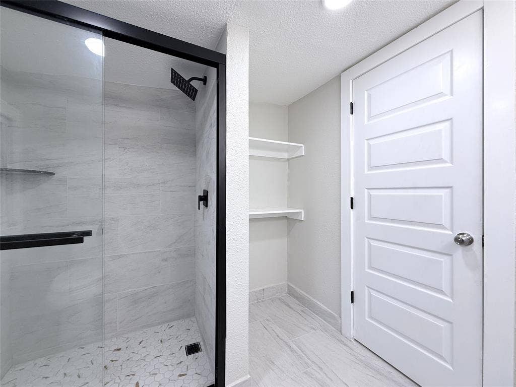 4522 Idaho Avenue, Dallas, TX 75216 Walk-in shower featuring matte black fixtures, a rainfall showerhead, glass enclosure, and light-toned tile surfaces, accompanied by built-in wall shelving and a five-panel interior door