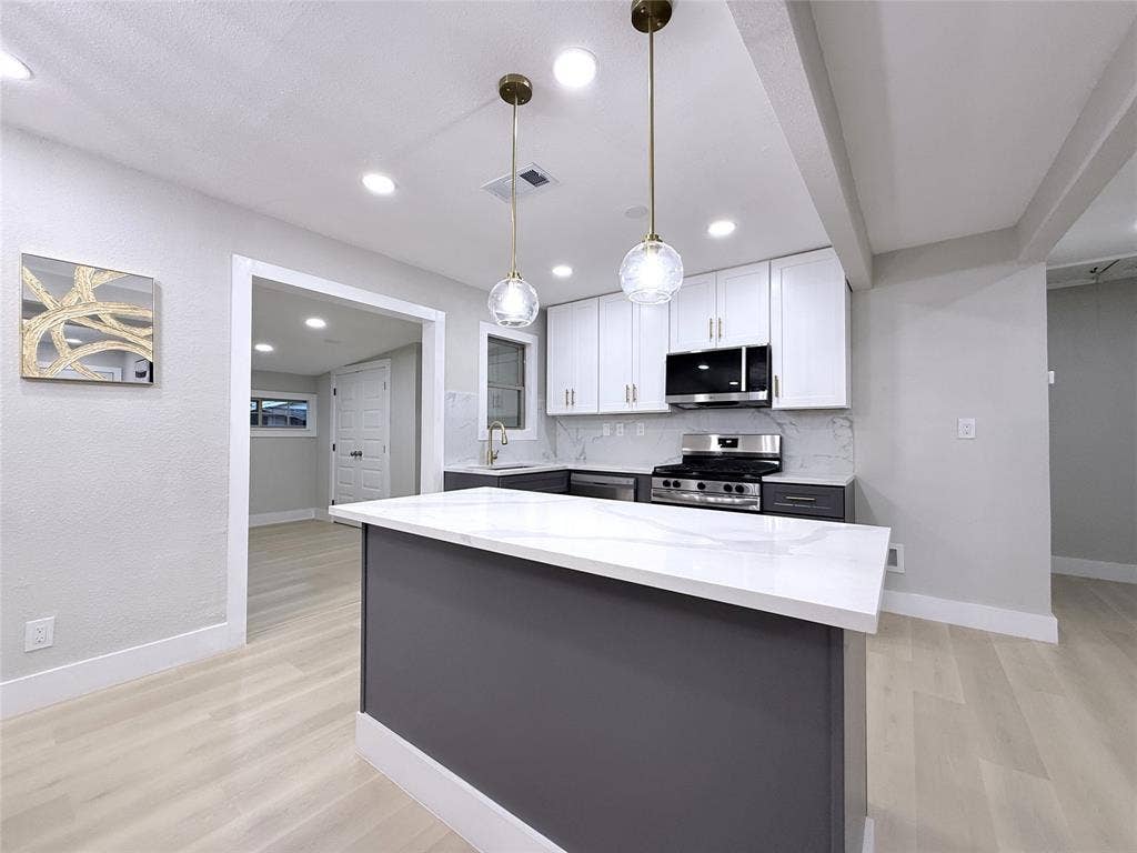 4522 Idaho Avenue, Dallas, TX 75216 Kitchen featuring a central island with a solid surface countertop, white shaker-style cabinetry, stainless steel appliances, and dual globe pendant lighting over the workspace