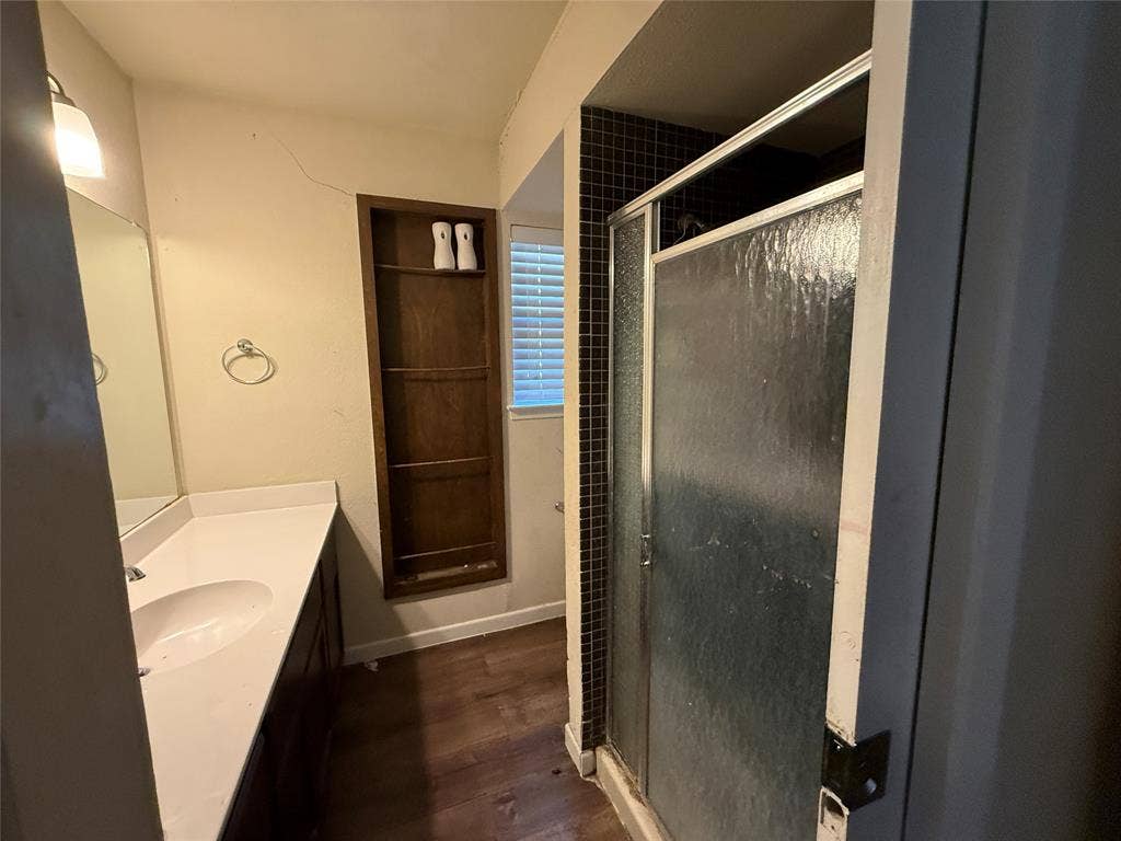 3825 Eugene Court N, Irving, TX 75062 Bathroom featuring wood-finish flooring, a vanity with a white countertop and oval under-mount sink, a recessed wood-finish shelving unit, a walk-in shower with dark tile surround and textured glass door, and a wall-mounted mirror