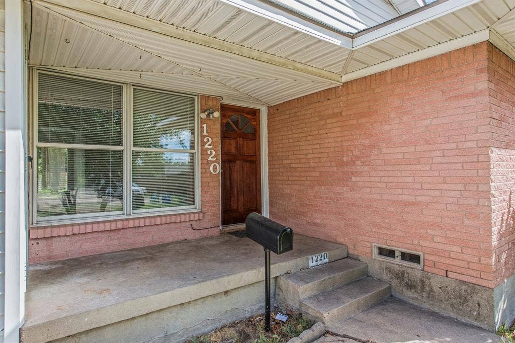 1220 Colfax Circle, Richardson, TX 75080 Brick facade exterior featuring a covered front porch, concrete steps, a wood-finish front door with arched glass inlay, and large multi-pane windows