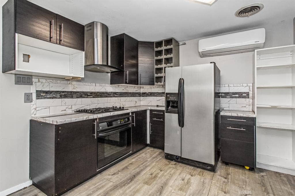 1220 Colfax Circle, Richardson, TX 75080 Dark-toned cabinetry featuring a built-in wine rack, stainless steel range hood, stone-patterned countertops, subway-style backsplash with decorative tile accents, and wood-finish flooring