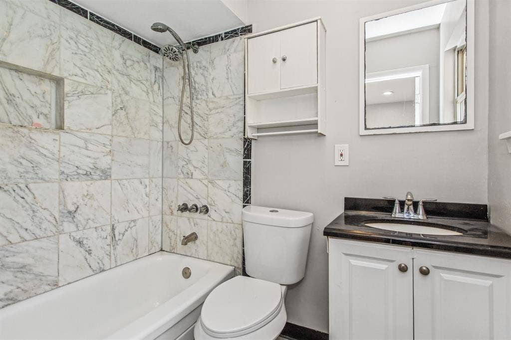 1220 Colfax Circle, Richardson, TX 75080 Bathroom featuring white cabinetry, dark stone vanity countertop with integrated oval sink, tub with stone-tiled surround, and wall-mounted storage cabinet