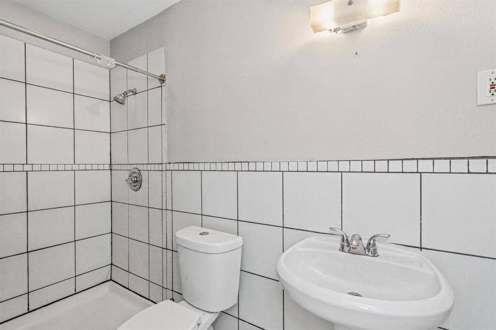 1220 Colfax Circle, Richardson, TX 75080 Bathroom featuring white square wall tiling with decorative mosaic accent border, pedestal sink with dual-handle chrome faucet, shower head with single-handle controls, and wall-mounted light fixture