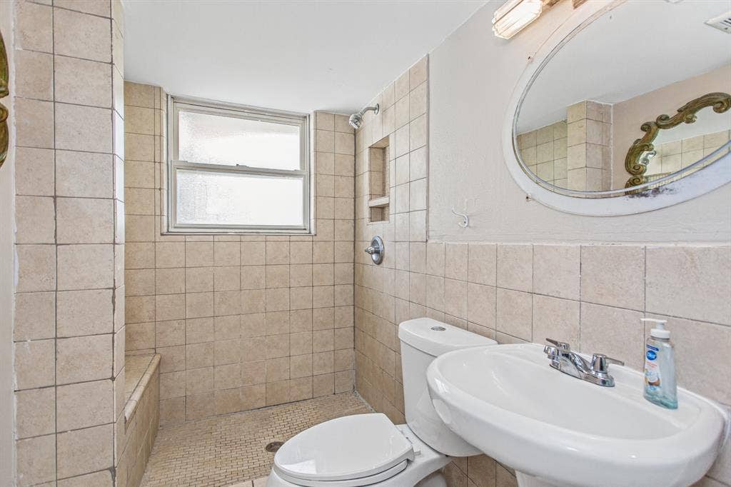 1220 Colfax Circle, Richardson, TX 75080 Full-tile shower surround with built-in wall niche, pedestal sink, chrome hardware, oval vanity mirror, and window providing natural illumination