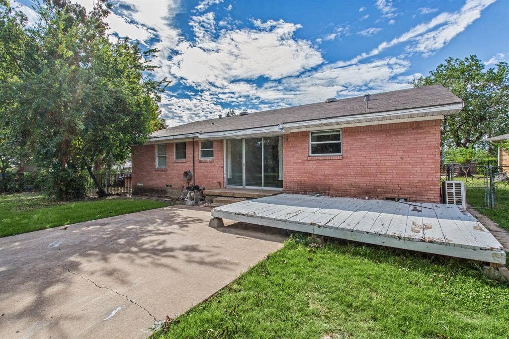 1220 Colfax Circle, Richardson, TX 75080 Exterior red brick facade featuring a sliding glass door, elevated wood deck, concrete patio, and fenced backyard with mature trees