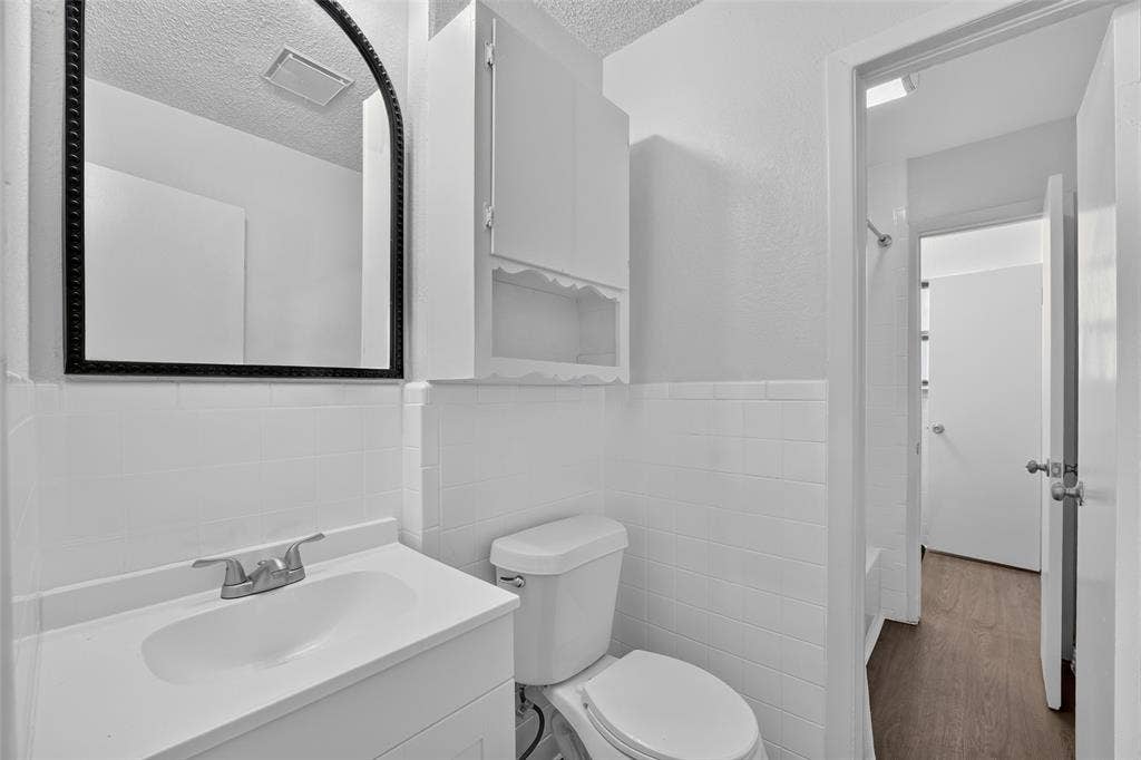 3532 Hilton Drive, Mesquite, TX 75150 White vanity with integrated sink, arched wall mirror with black trim, built-in upper cabinet, white subway tile wall surround, and wood-finish flooring