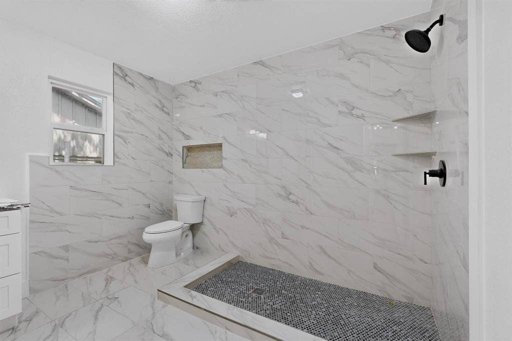 550 Glen Oaks Boulevard, Dallas, TX 75232 Walk-in shower featuring white large-format stone-patterned tile, matte black rainfall showerhead, integrated recessed wall niche, and dark pebble-stone flooring
