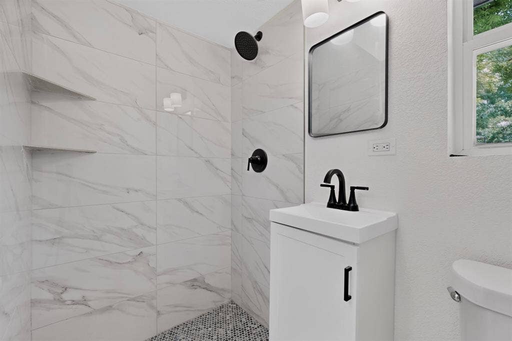 550 Glen Oaks Boulevard, Dallas, TX 75232 Shower featuring large-format veined tile walls and a built-in shelf, complemented by a pedestal vanity with a matte black faucet and a wall-mounted mirror