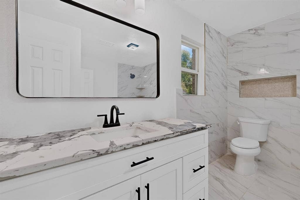 550 Glen Oaks Boulevard, Dallas, TX 75232 Bathroom featuring a white vanity with stone-finish countertop, undermount sink with matte black widespread faucet, rectangular mirror with black frame, and tiled walk-in shower with recessed niche