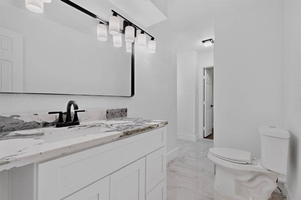 550 Glen Oaks Boulevard, Dallas, TX 75232 White cabinetry with stone-finish countertop, undermount sink, matte black faucet, six-light vanity fixture, and large rectangular mirror