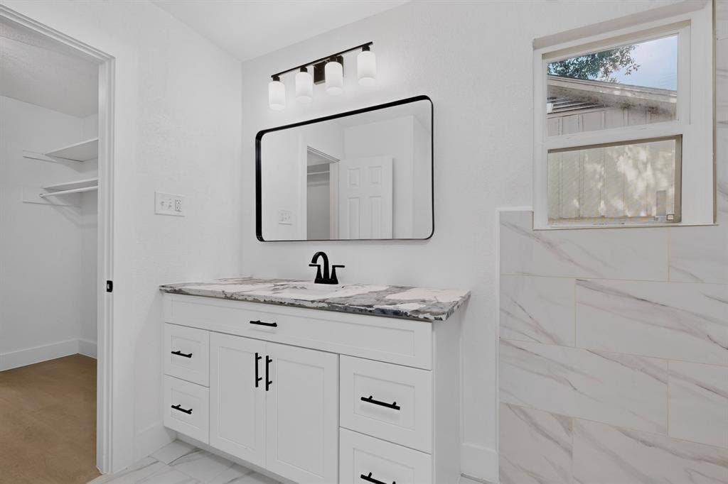 550 Glen Oaks Boulevard, Dallas, TX 75232 White vanity with stone-patterned countertop, rectangular undermount sink, black matte hardware, modern wall-mounted light fixture, and framed mirror