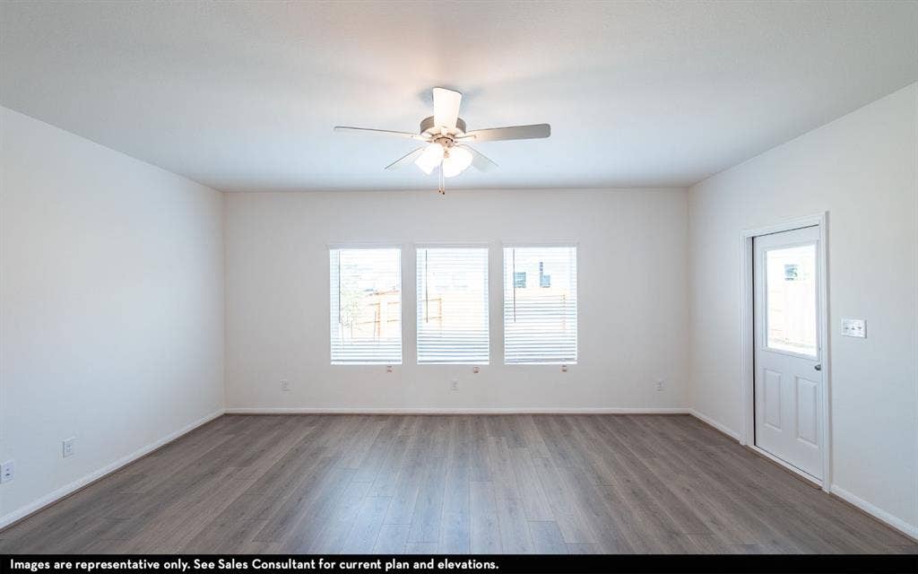 Neutral-toned interior space featuring wood-finish flooring, three windows with integrated blinds, a central ceiling fan with multi-light fixture, and a glass-paneled exterior door