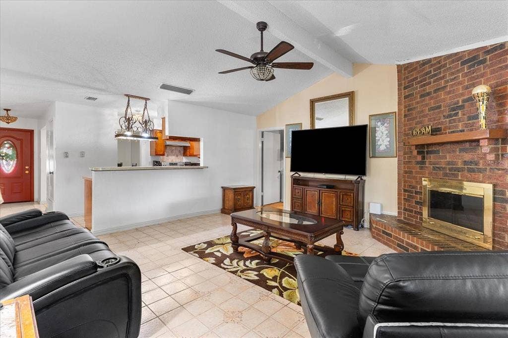 2702 Pebble Beach Drive, Lancaster, TX 75134 Living area featuring a vaulted ceiling with a wooden fan, floor-to-ceiling brick fireplace with a wood mantel, tiled flooring, and a pass-through opening to the kitchen