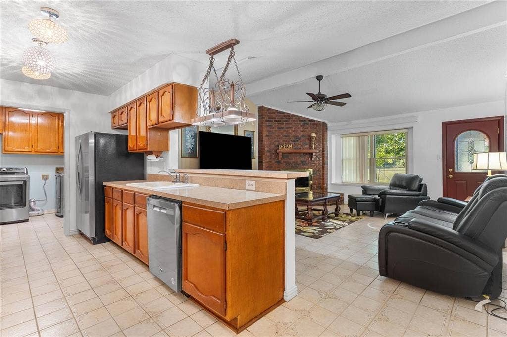 2702 Pebble Beach Drive, Lancaster, TX 75134 Kitchen featuring wooden cabinetry, a peninsula countertop, and integrated stainless steel dishwasher, with a brick fireplace and decorative pendant lighting