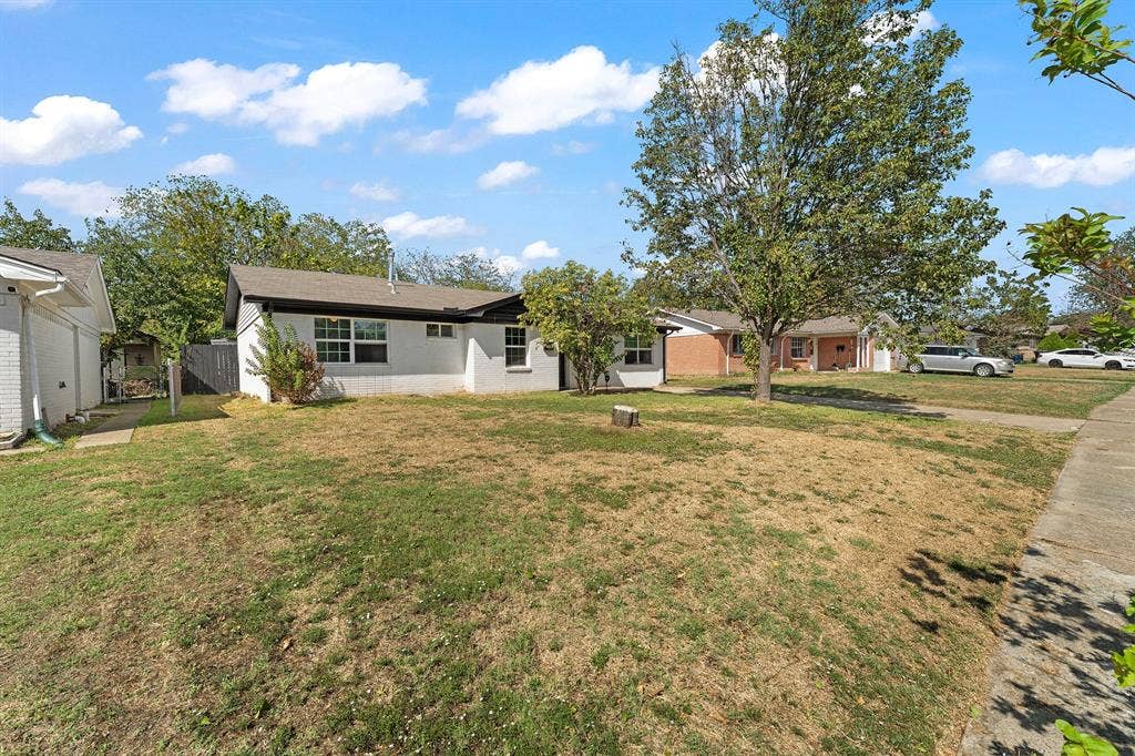 Single-story residence featuring white painted brick siding, a neutral-toned shingled roof, grid-pattern windows, and a generous front lawn with mature shade trees