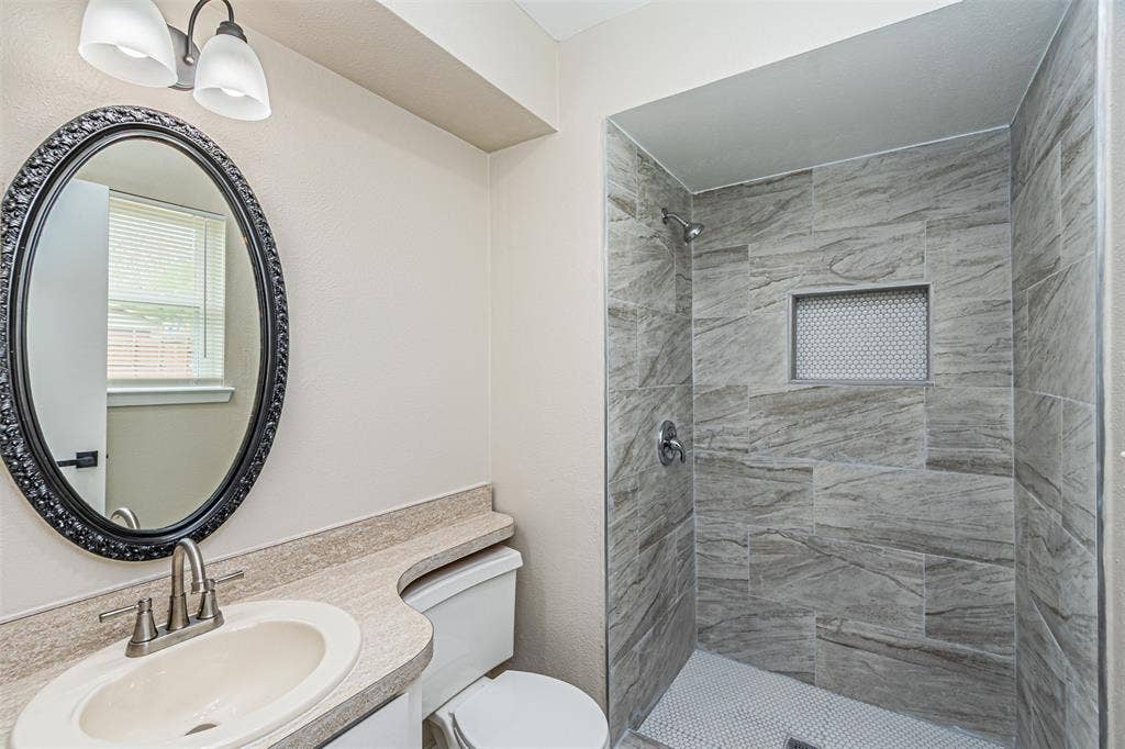 1509 Crockett Street, Garland, TX 75042 Bathroom featuring a walk-in shower with stone-patterned wall tile, a recessed niche, a neutral-toned countertop with an integrated basin, and an oval mirror with a decorative frame