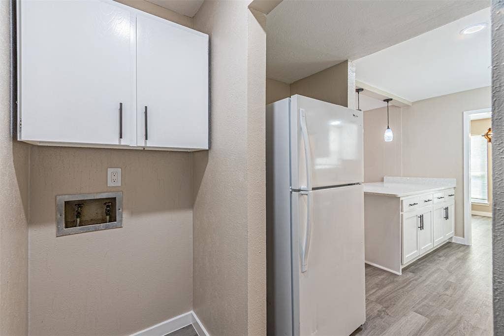 1509 Crockett Street, Garland, TX 75042 Dedicated utility area featuring white wall-mounted cabinetry and recessed plumbing connections, paired with a white refrigerator and kitchen cabinetry with light-toned countertops, finished with wood-finish flooring