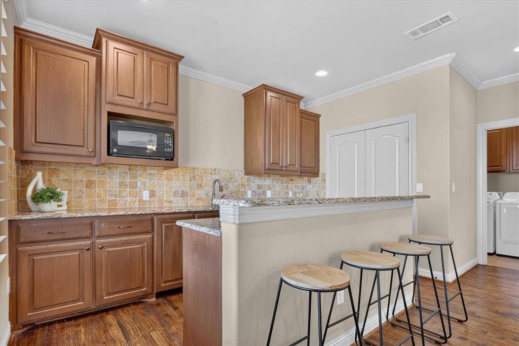 563 Rockingham Drive, Irving, TX 75063 Wet bar kitchen featuring wood cabinetry, granite countertops, square-tile backsplash, built-in microwave, mini sink, and mini fridge. Handscraped wood flooring throughout.