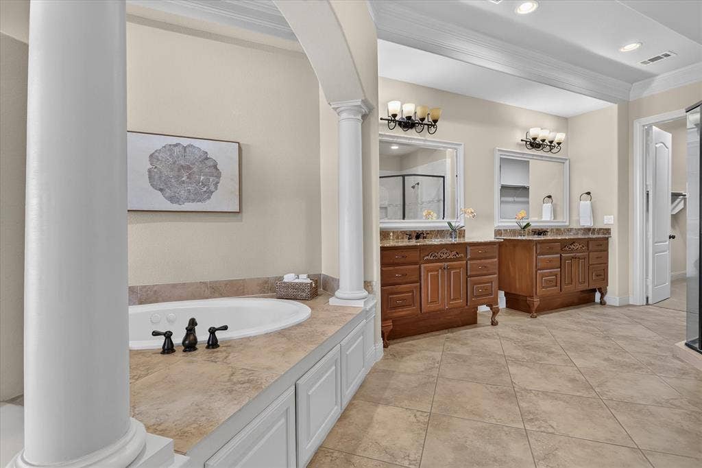 563 Rockingham Drive, Irving, TX 75063 Bathroom featuring dual wood cabinetry vanities, stone-finish countertops, tiled soaking tub surround with decorative columns, and tile flooring