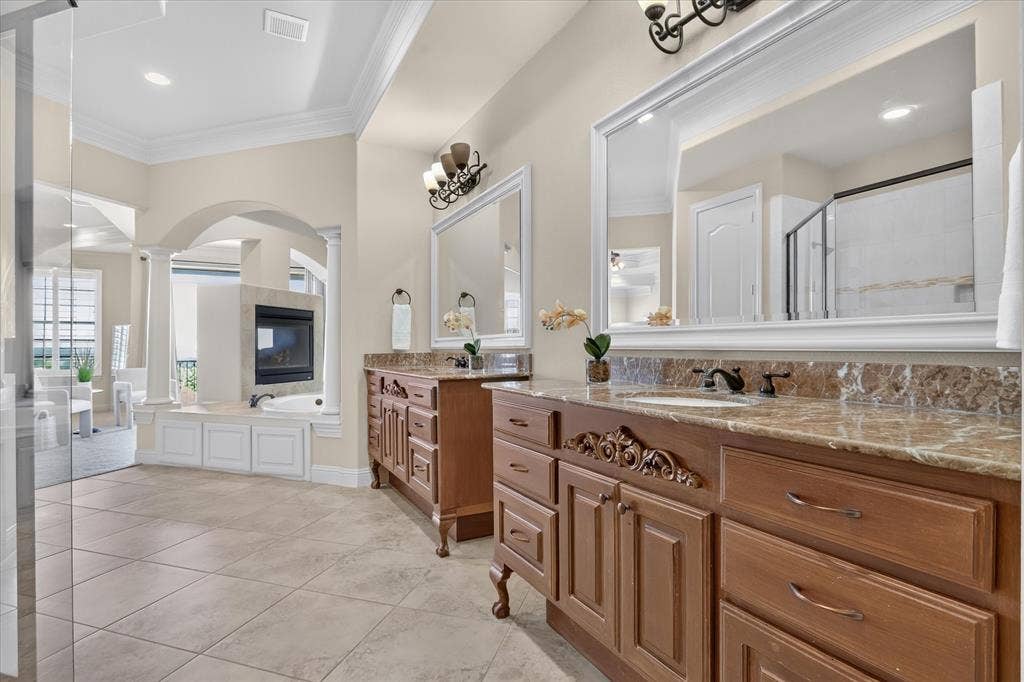 563 Rockingham Drive, Irving, TX 75063 Dual wood vanities featuring stone countertops, large framed wall mirrors, a deep soaking tub, a built-in fireplace, and tile flooring