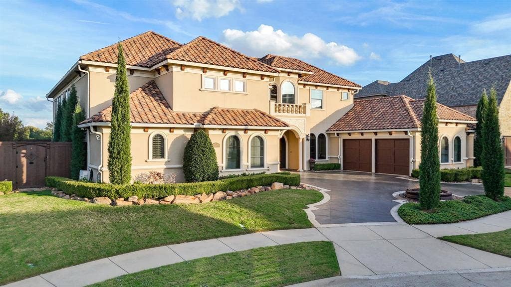 6590 Gerrard Street, Frisco, TX 75034 Two-story stucco exterior featuring a multi-gabled tile roof, arched windows, a stone-bordered front garden, and a concrete driveway leading to a three-car garage