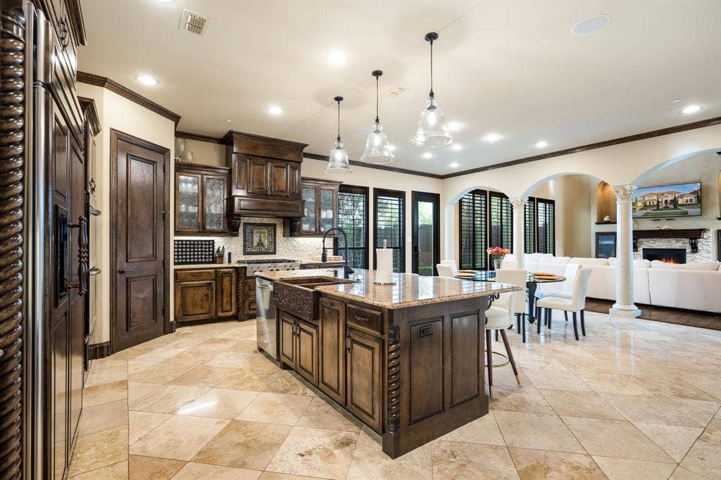 6590 Gerrard Street, Frisco, TX 75034 Kitchen featuring custom wood cabinetry, an expansive center island with a farmhouse sink, stone countertops, stone flooring, and decorative column accents
