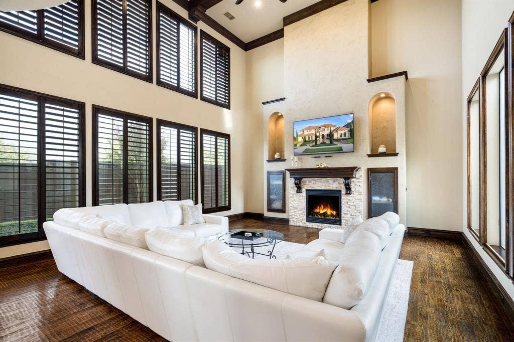 6590 Gerrard Street, Frisco, TX 75034 Two-story living area featuring wood-finish flooring, floor-to-ceiling windows with dark-finish shutters, a stacked stone fireplace with a wood mantel, and custom built-in arched alcoves