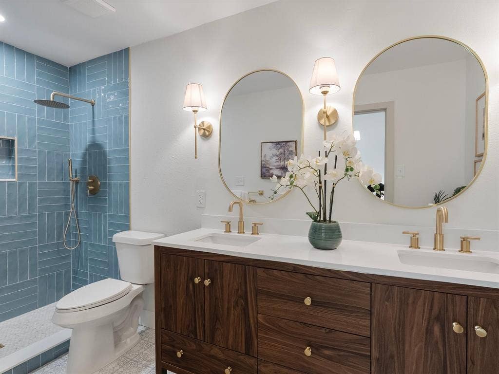 Double vanity featuring a wood-finish cabinet, white solid-surface countertop, dual integrated sinks, gold-tone hardware, and oval-shaped gold-rimmed mirrors with wall-mounted sconces