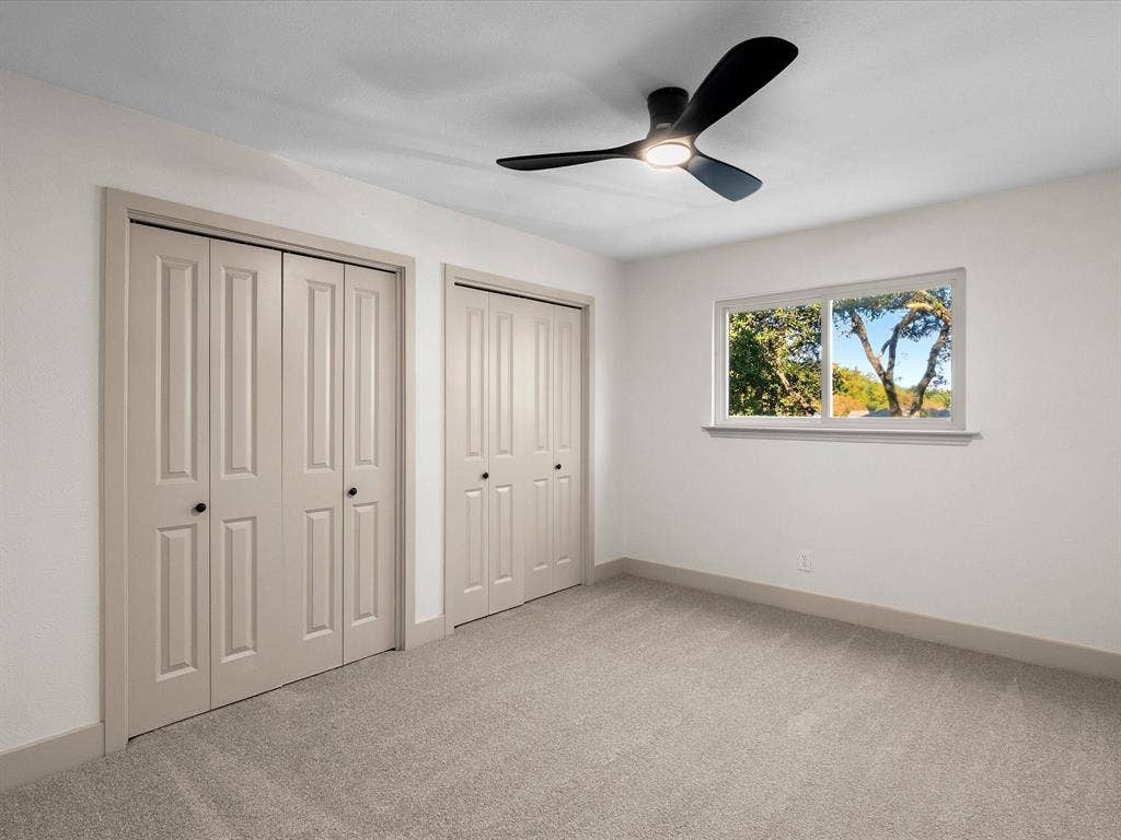 Neutral-toned interior featuring dual bi-fold closets, carpeted flooring, a horizontal sliding window, and a matte black three-blade ceiling fan with integrated lighting