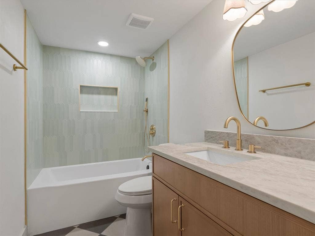 Bathroom featuring a wood-finish vanity with a stone-top counter, undermount sink, brass-tone hardware, oval mirror, and a tiled shower surround with a built-in niche