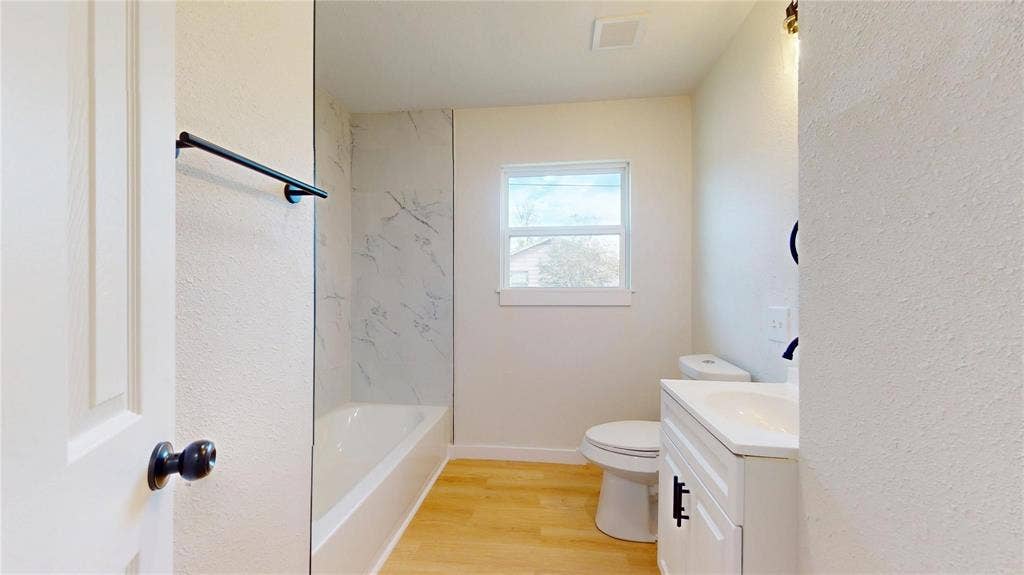3901 Park Street, Greenville, TX 75401 Bathroom featuring wood-finish flooring, a white vanity with an integrated countertop, matte black hardware, and a tub enclosure with light-toned patterned wall surfacing