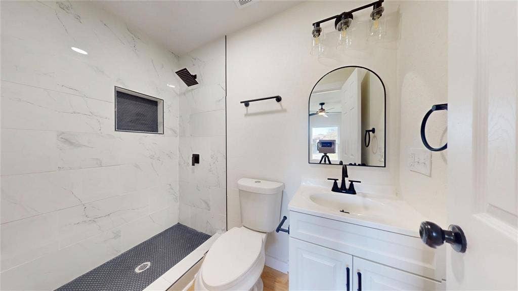 3901 Park Street, Greenville, TX 75401 Walk-in shower with floor-to-ceiling stone-pattern tile, contrasting dark mosaic floor, built-in recessed wall niche, arched vanity mirror, and matte black hardware