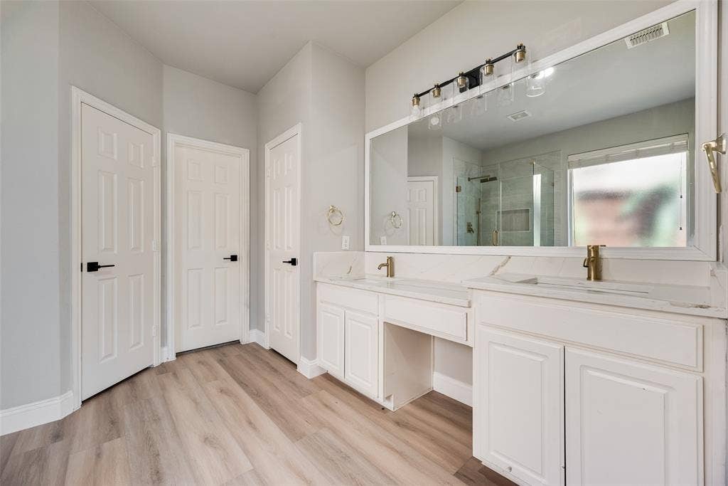1716 Clarke Springs Drive, Allen, TX 75002 Dual-sink vanity with white cabinetry, stone-finish countertops, brass-toned fixtures, and a wide-frame wall mirror, complemented by wood-finish flooring
