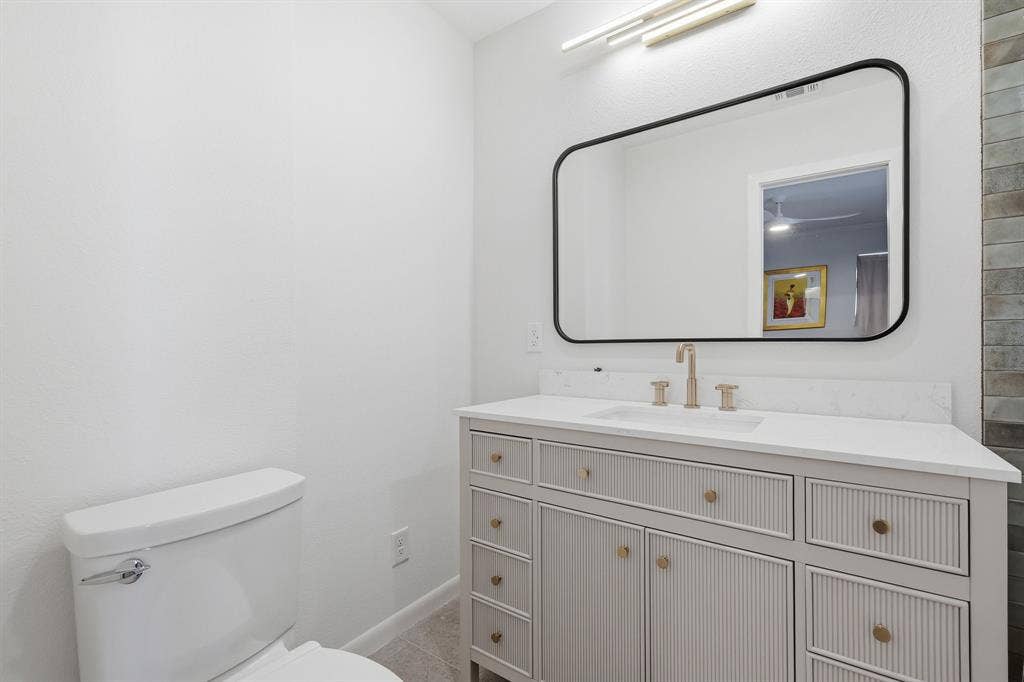 2810 Bonnie View Road, Dallas, TX 75216 Vanity featuring reeded-texture cabinetry, stone-surface countertop, gold-tone hardware, rectangular undermount sink, and wall-mounted gold-tone faucet, accompanied by an arched black-framed mirror and linear LED light fixture