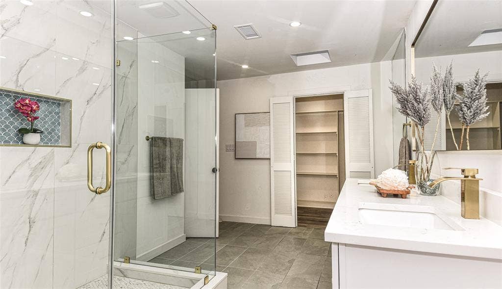 13936 Tanglewood Place, Farmers Branch, TX 75234 Ensuite Bath to the Primary Bedroom has it all...Walk-in shower with floor-to-ceiling veined tile, glass enclosure, gold-toned hardware, and decorative niche, paired with a dual-sink vanity featuring stone countertops and gold-toned fixtures