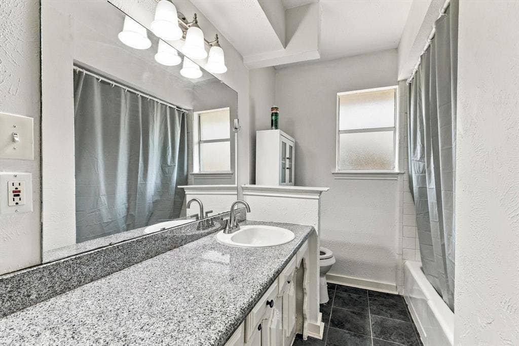 1900 Ports O Call Drive, Plano, TX 75075 Full-width vanity featuring a stone countertop, integrated sink, large wall-mounted mirror, and multi-bulb vanity light fixture, complemented by dark tile flooring and a tub-shower combination