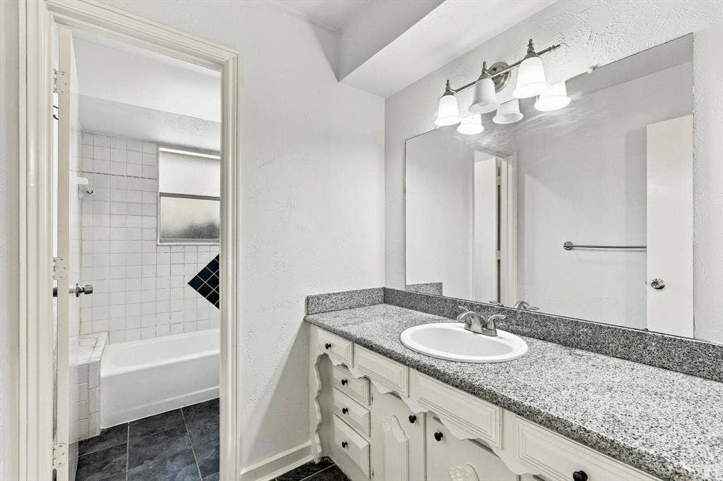 1900 Ports O Call Drive, Plano, TX 75075 Bathroom featuring a vanity with stone-finish countertop, oval undermount sink, expansive wall mirror, and five-light vanity fixture, including a tile-surround bathtub and dark tile flooring