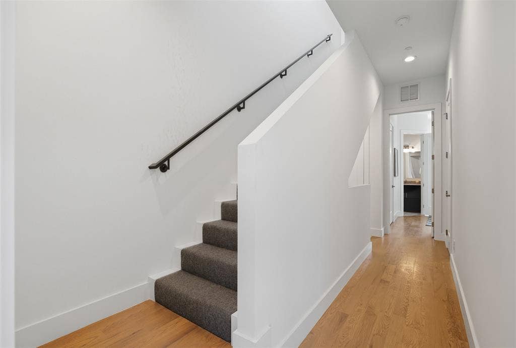 Staircase featuring charcoal carpeted treads, a solid half-wall, and a matte black wall-mounted handrail, complemented by wood-finish flooring and recessed lighting