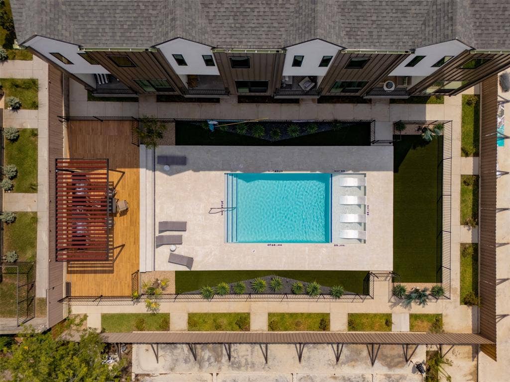 Outdoor area featuring a swimming pool with a built-in tanning ledge, wood-finish decking, a fixed slatted pergola, and artificial turf lawn areas enclosed by perimeter fencing