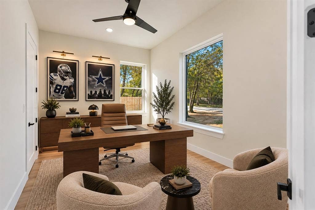 Neutral-toned room featuring wood-finish flooring, two large windows, a matte black ceiling fan with integrated lighting, and dual wall-mounted picture lights