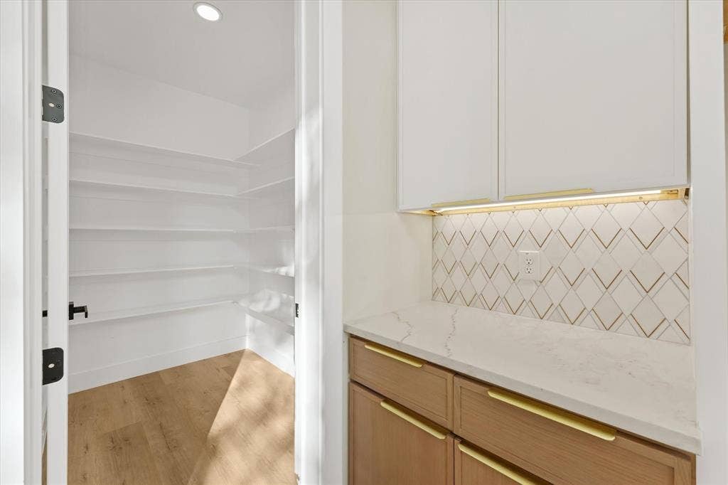 Custom cabinetry featuring light-toned wood base units, white upper cabinets, stone-composite countertops, geometric tile backsplash, and wood-finish flooring