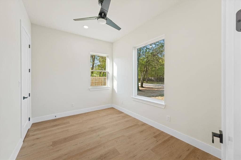 Neutral-toned interior featuring wood-finish flooring, a matte black ceiling fan, recessed lighting, and large windows with exterior foliage