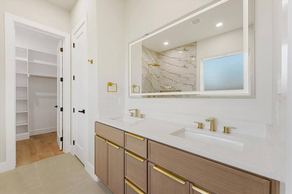 Dual-sink vanity with a quartz countertop, gold-toned hardware, illuminated mirror, and integrated shelving in the walk-in closet