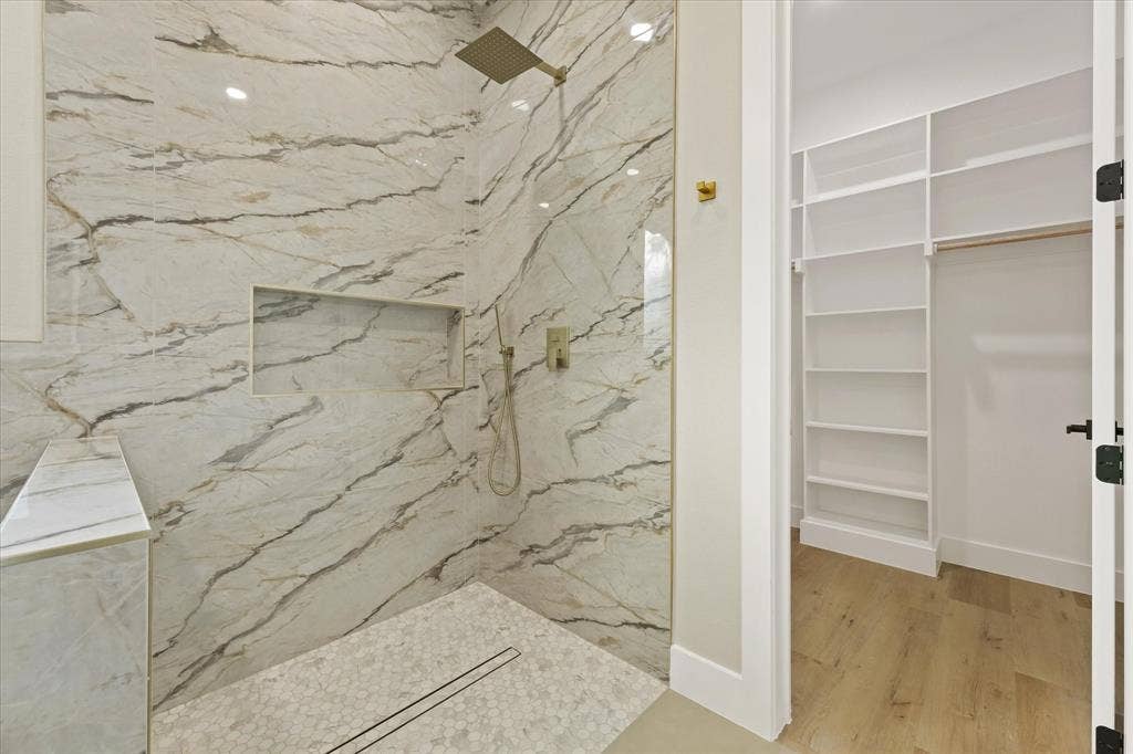 Walk-in shower featuring floor-to-ceiling stone-patterned wall tile, a built-in recessed niche, a rainfall showerhead, and a stone-tiled floor with a linear drain, plus a custom built-in closet with shelving and wood-finish flooring