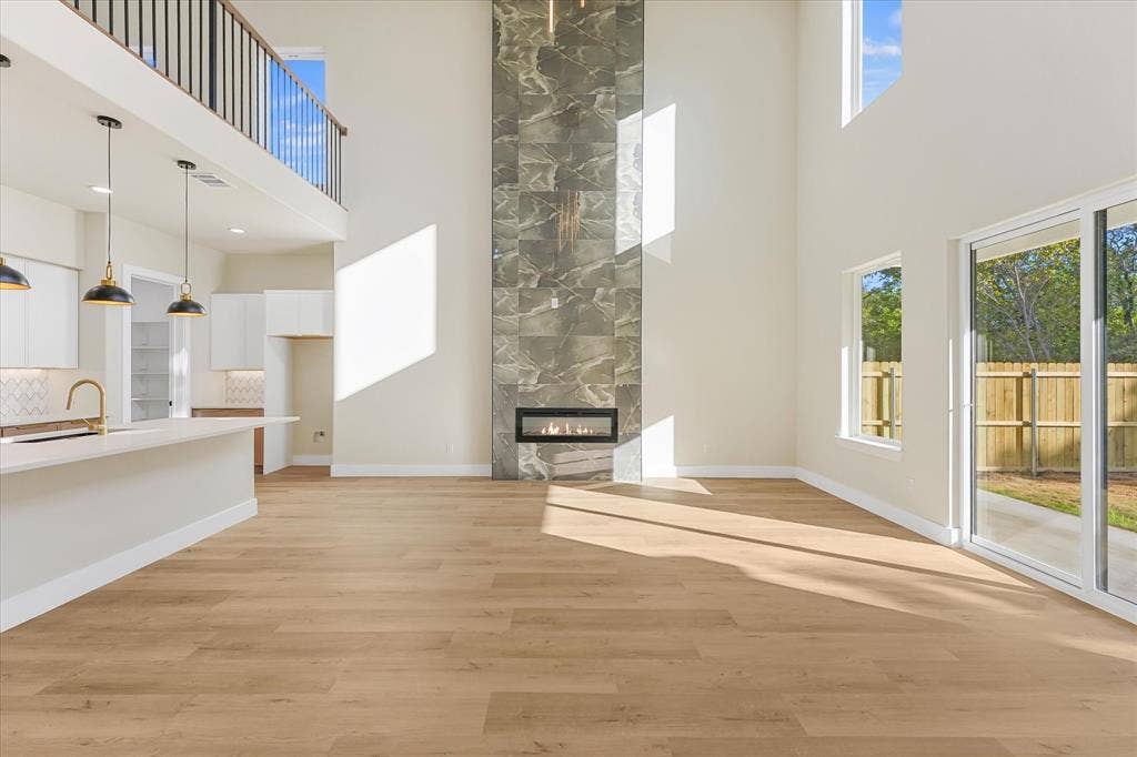 Double-height living area featuring a full-height stone-clad fireplace, wood-finish flooring, white cabinetry with a quartz countertop island, pendant lighting, and floor-to-ceiling windows