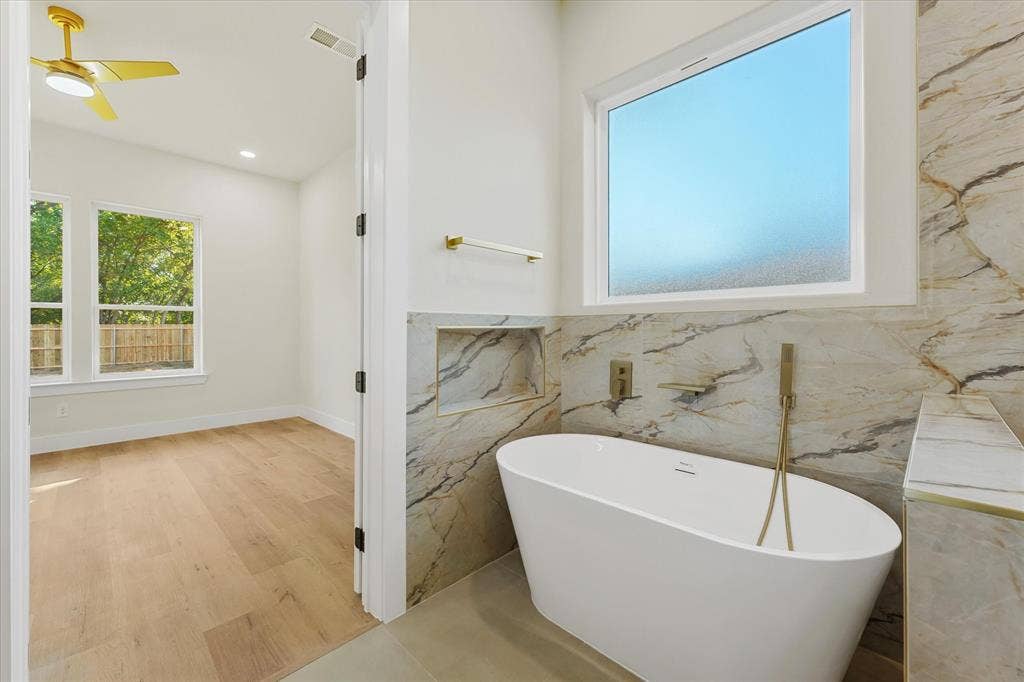 Freestanding soaking tub with a wall-mounted brass hand shower, floor-to-ceiling stone-patterned tile surround, built-in recessed wall niche, and wide-plank wood-finish flooring