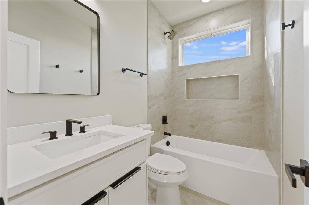 Bathroom featuring a white vanity with a solid-surface countertop, rectangular undermount sink, matte black hardware, and a tub-shower combination with tiled wall surround and recessed shelving