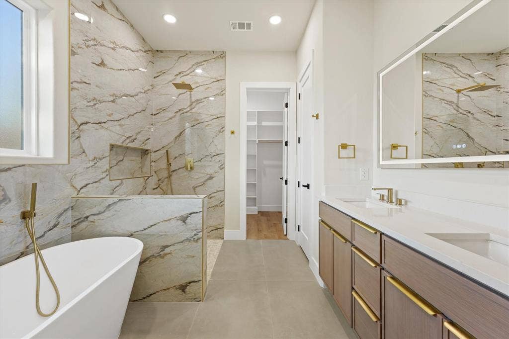 Freestanding soaking tub with a wall-mounted brass handheld faucet, walk-in shower featuring large-format decorative tile surround, dual-sink vanity with quartz countertops and brass hardware, and a lighted vanity mirror