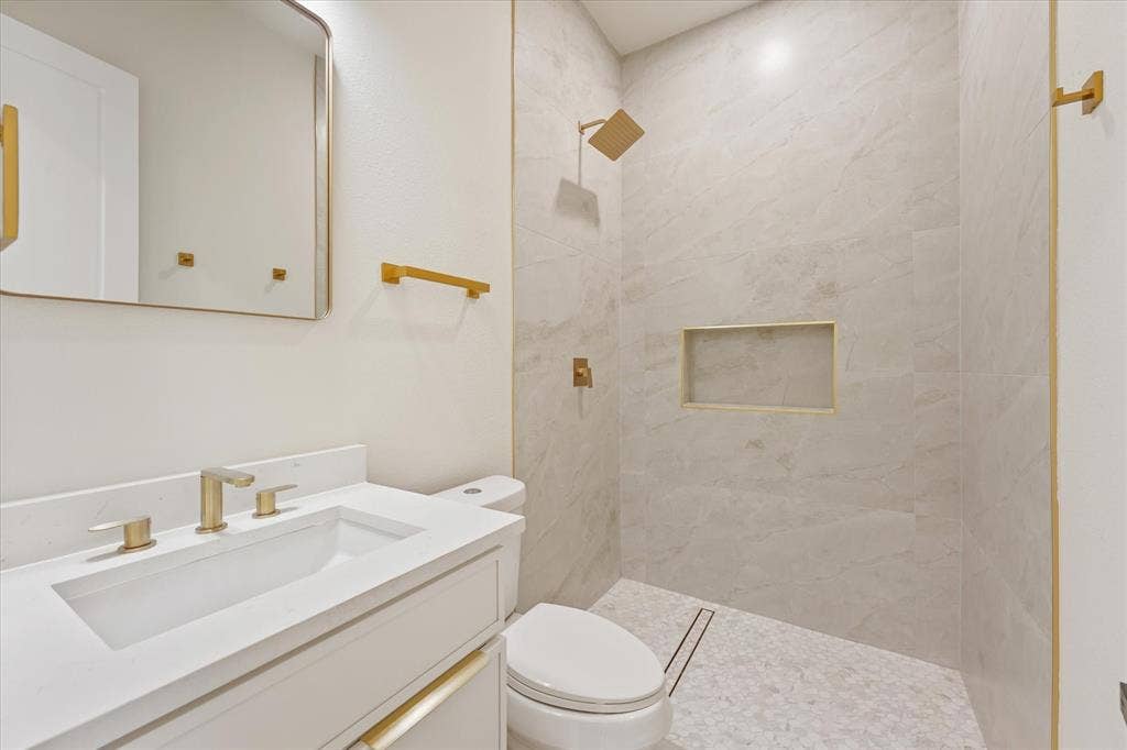 Bathroom featuring a white vanity with a rectangular sink, stone-finish countertop, walk-in shower with floor-to-ceiling tile, built-in wall niche, and gold-tone hardware